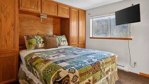 Un dormitorio con una cama grande, armarios de madera, un televisor montado y una ventana con vista a árboles.