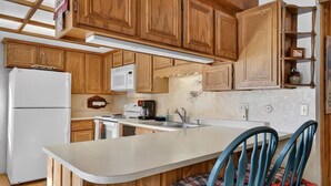 Cocina con armarios de madera, un refrigerador blanco, una cafetera y dos sillas azules con cojines a cuadros.