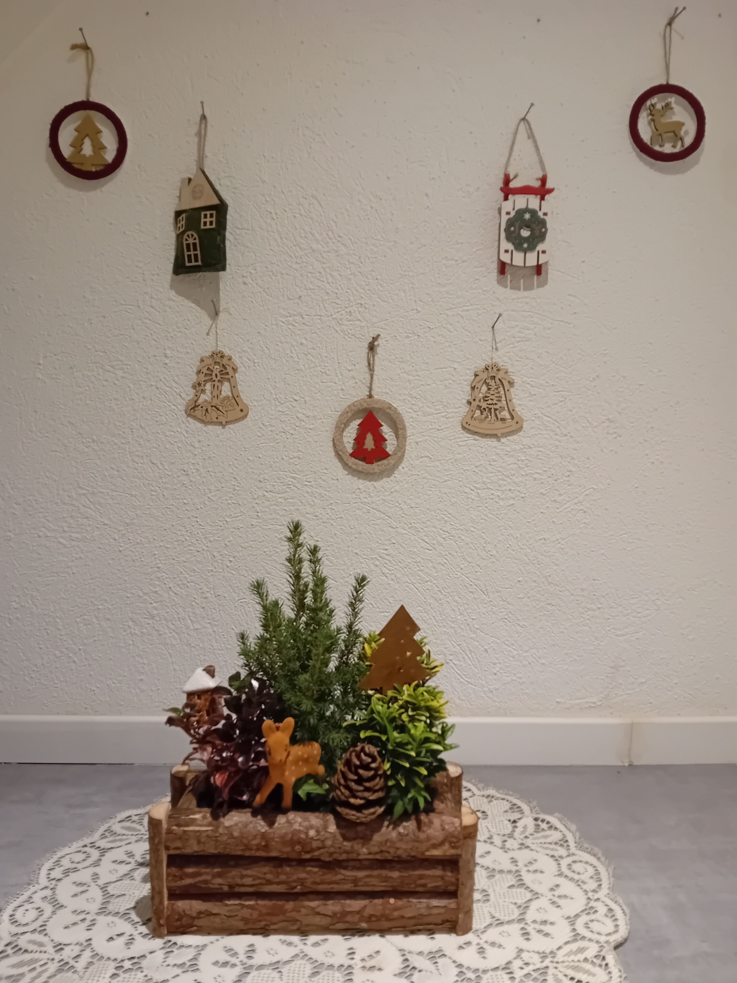 Un pot de fleurs en bois, orné d’un sapin de Noël, d’une pomme de pin et d’objets décoratifs, le tout posé sur un chemin de table en dentelle.