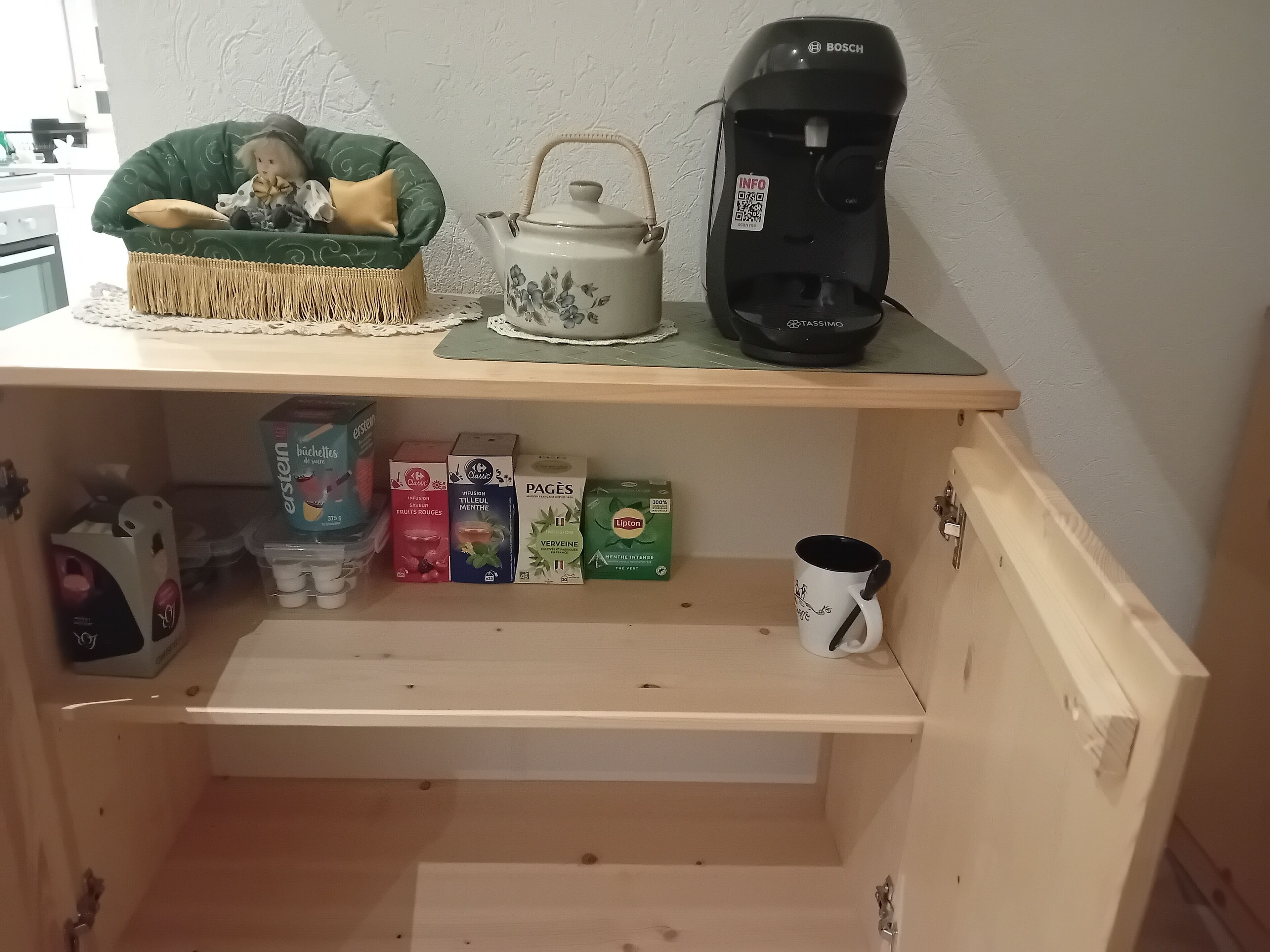 Una kitchenette con cafetera, tetera y diversos alimentos en los estantes.
