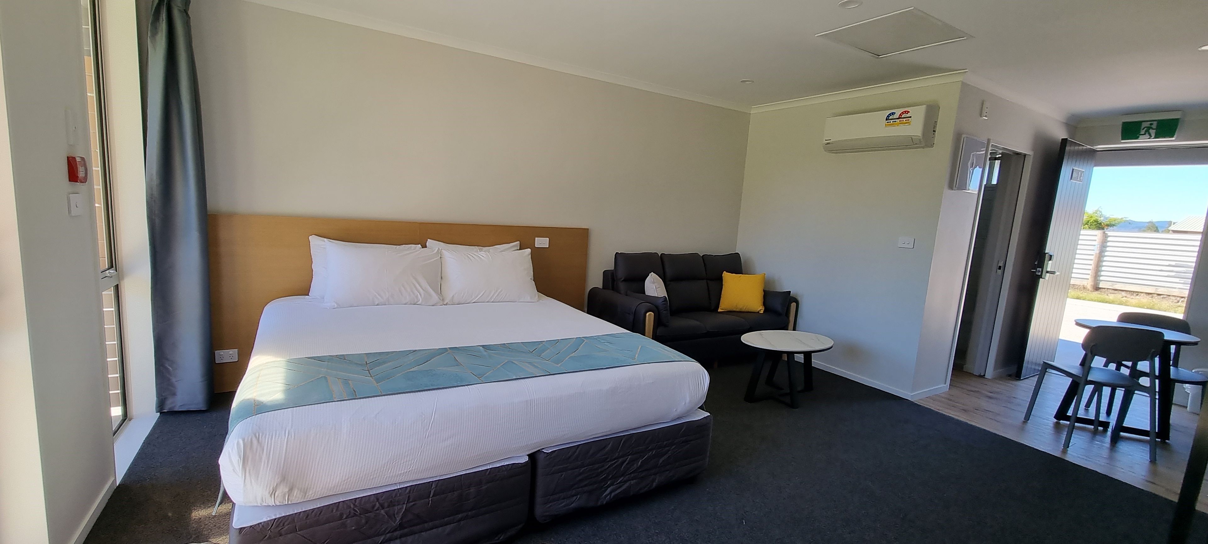 A hotel room with a large bed, a sofa, a small round table, and an air conditioning unit on the wall.