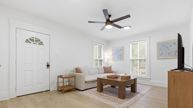 A living room with a white door, a ceiling fan, a white sofa, a coffee table, and a television on a wooden stand. - Charming 2BR Retreat in North Little Rock, Close to Hospitals & Downtown (North Little Rock)