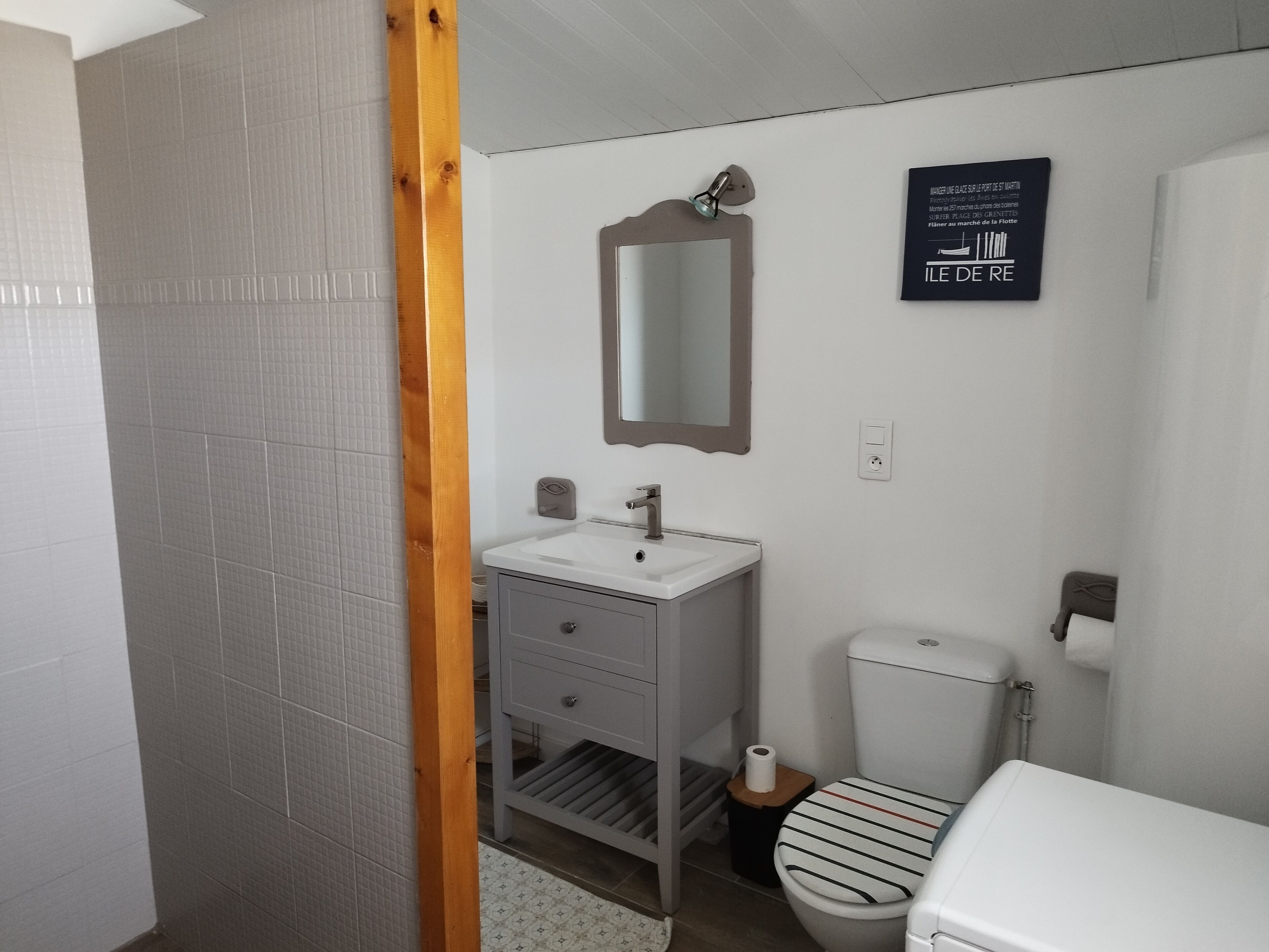 Ein Badezimmer mit einem weißen Waschbecken, Spiegel und WC. Ein Holzrahmen an der Tür und ein Schild an der Wand sind ebenfalls zu sehen.
