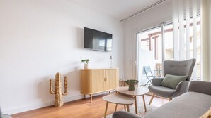 Ein modernes Wohnzimmer mit einem Flachbildfernseher, einer Holzkabine, einem Kaktus, einem grauen Sessel und einem kleinen Tisch.