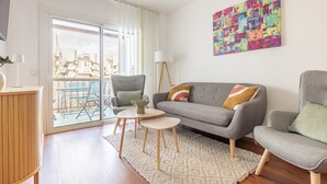 Ein modernes Wohnzimmer mit grauem Sofa, zwei Sesseln, einem kleinen Couchtisch und einer Stehlampe. Ein farbenfrohes abstraktes Gemälde hängt an der Wand, und durch eine Schiebetür ist ein Blick auf Gebäude möglich.
