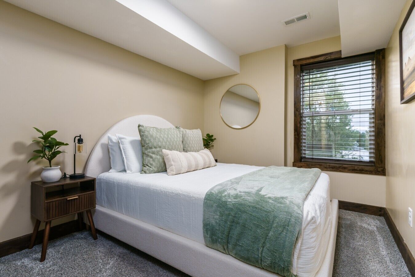Une chambre à coucher avec un lit, une table de chevet, un miroir, une fenêtre et une plante.