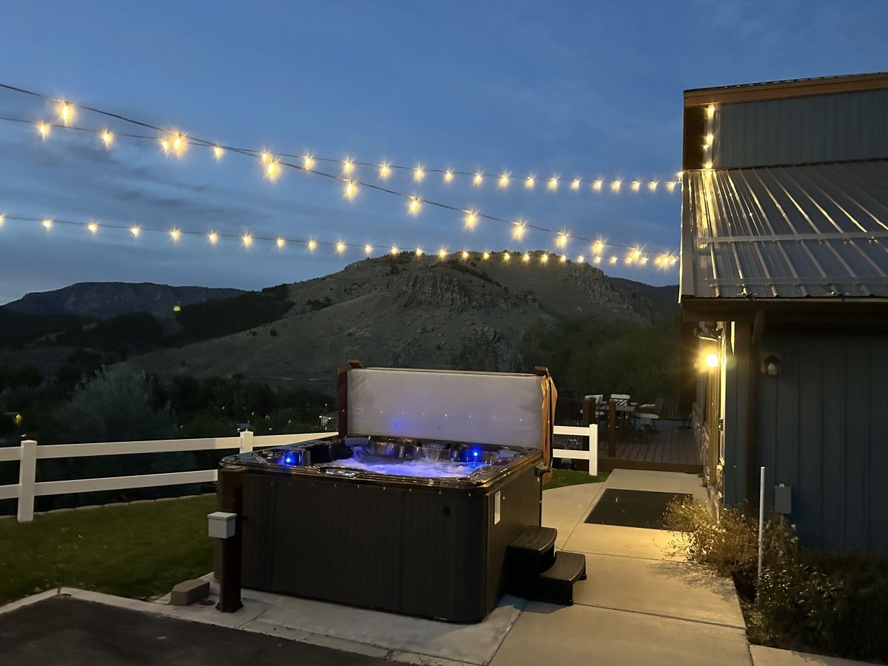 Un jacuzzi extérieur avec une terrasse en bois, entouré d’une clôture blanche et illuminé de guirlandes lumineuses.