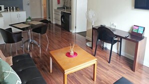 Een moderne woonkamer met een houten salontafel, zwarte stoelen, een bank, een eettafel met stoelen, een keuken met een wasmachine, en een aan de muur bevestigde televisie.