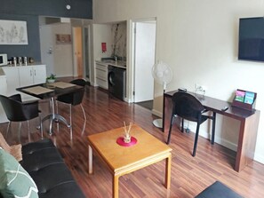 Ein modernes Wohnzimmer mit Couch, schwarzen Sesseln, einem Holztisch, einem Esstisch mit Stühlen, einer Küchenzeile inklusive Waschmaschine und einem an der Wand befestigten Fernseher.