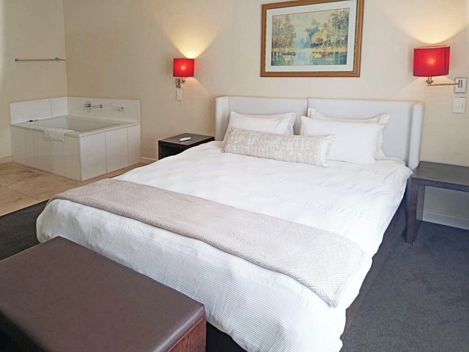 Une chambre d’hôtel avec un grand lit, une table de chevet, une commode, une petite table, un banc, une baignoire et un tableau au mur.