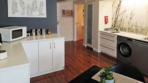 Een moderne keuken met witte kastjes, een magnetron en een wasmachine. Er is een eethoek met een tafel en stoelen, en een plant op de tafel.