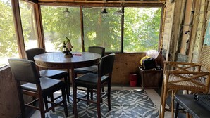 Una habitación con una mesa redonda de madera, cuatro sillas, un tapete estampado, una ventana con vistas a follaje y una silla de mimbre.