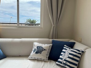 A room with a view of the beach through a window, a beige sofa with two pillows, one with a fish design and another with a striped pattern. - Palm Garden Apartment with terrace, amazing ocean view and swimming pool. (Morro Jable)
