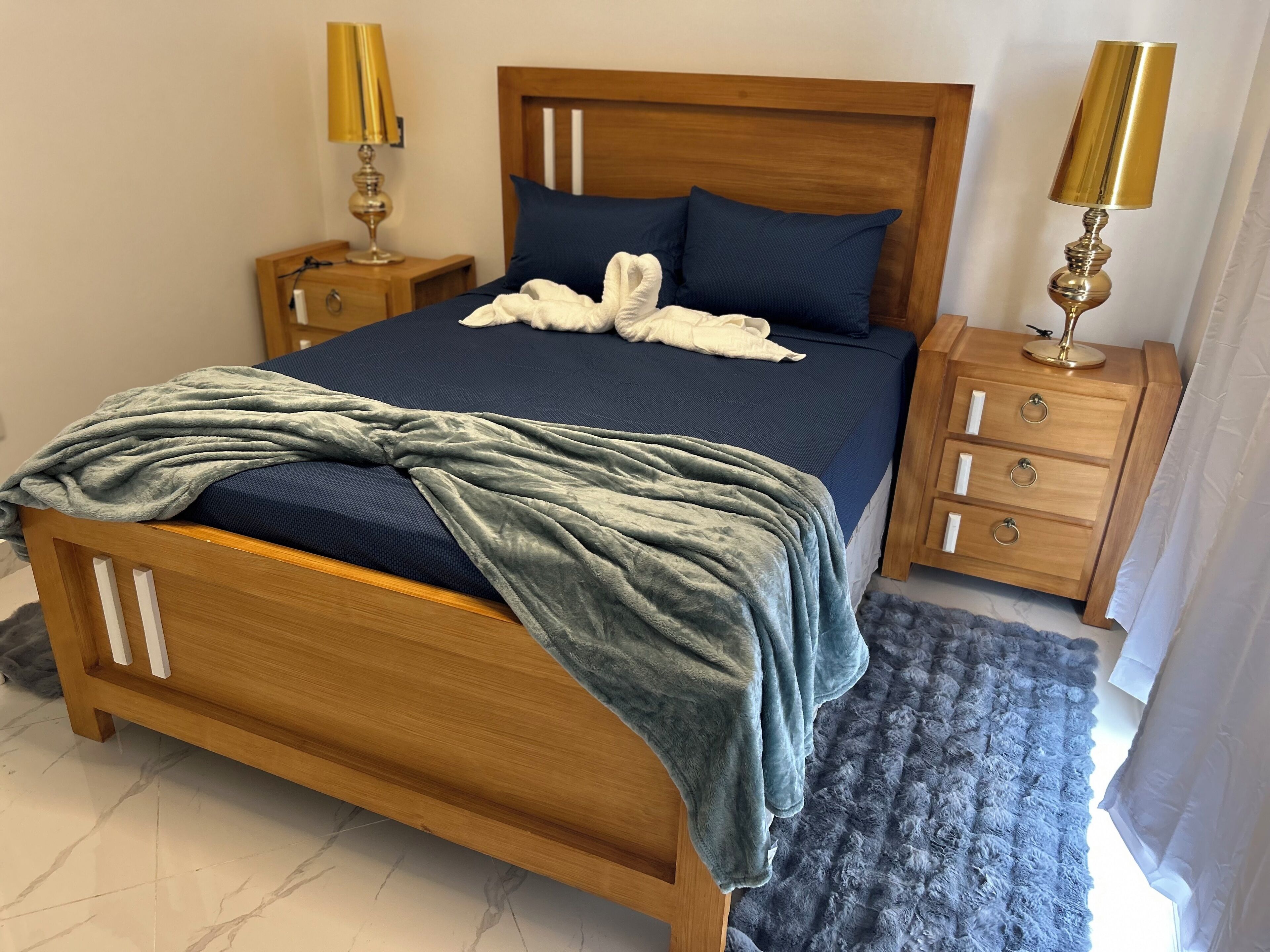 Una cama bien hecha con edredón azul y manta gris, cabecera de madera, dos mesitas de noche con lámparas y una alfombra gris.