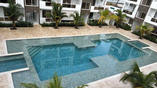 Ein Pool mit geometrischem Design, umgeben von einer angelegten Fläche mit Palmen und einem modernen Gebäude im Hintergrund.