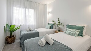 Ein Hotelzimmer mit zwei Betten, einem großen Fenster mit durchsichtigen Vorhängen, einer Zimmerpflanze und einer Wandleuchte.