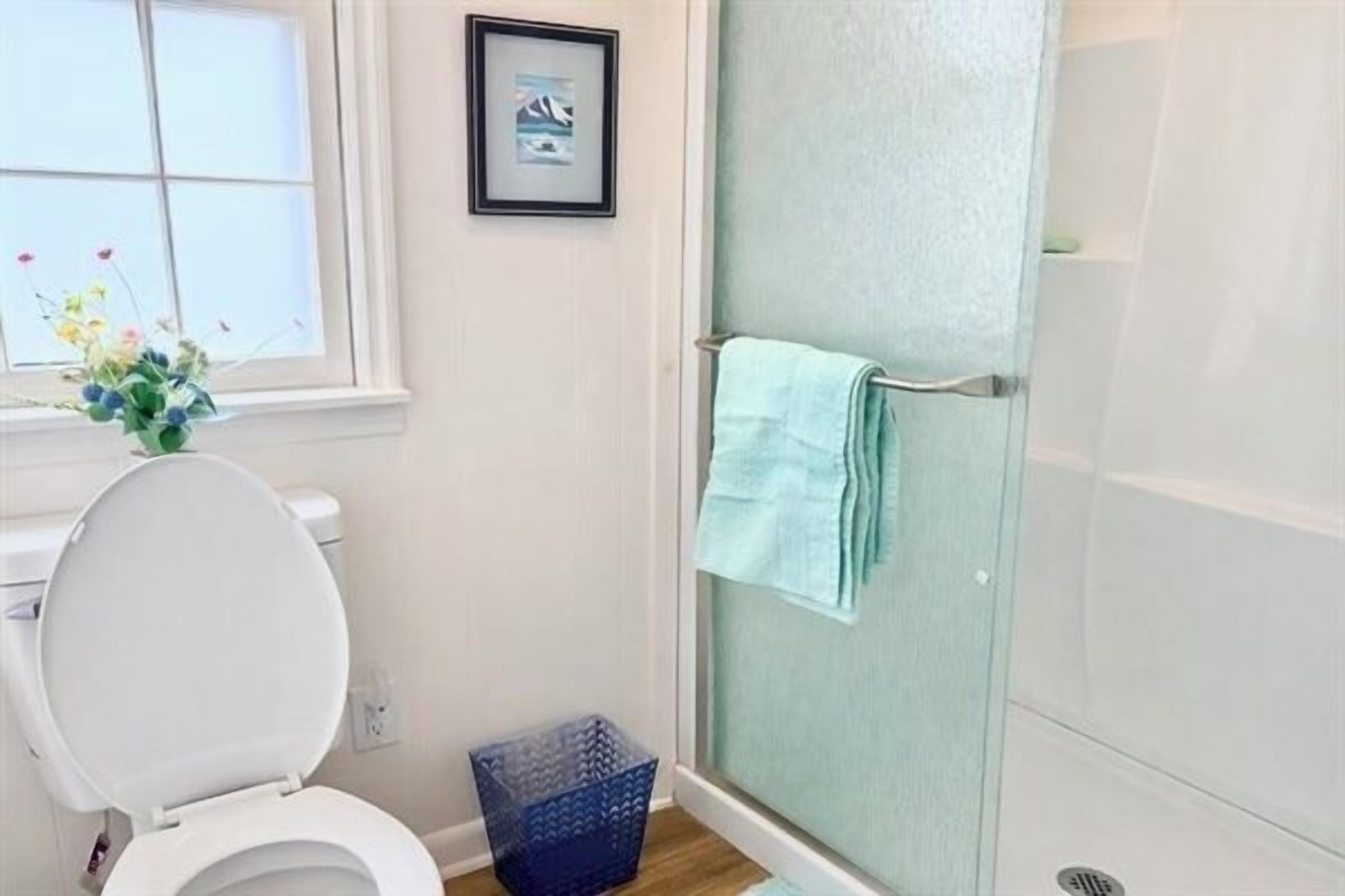 Un baño con inodoro, un cuadro en la pared, ducha con toalla y una cesta azul.