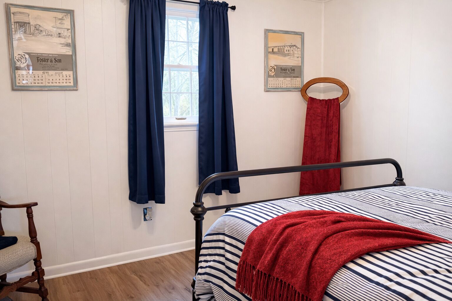 Quarto com cama, cadeira de madeira, janela com cortinas e duas imagens emolduradas na parede.