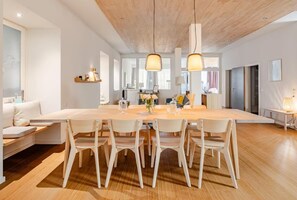 Een moderne eetruimte met een lange houten tafel, witte stoelen, hanglampen en een keuken op de achtergrond.