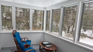 A corner room with a rocking chair, a small table, and large windows with a view of trees.