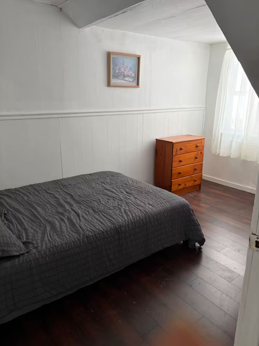 Een slaapkamer met een bed, een houten dressoir, een lijst met een afbeelding aan de muur, en een raam met gordijnen.