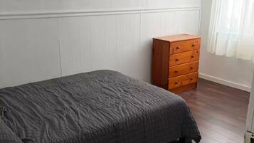 Een slaapkamer met een bed, een houten dressoir, een lijst met een afbeelding aan de muur, en een raam met gordijnen.