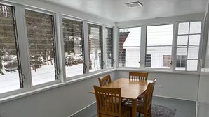 A dining area with a wooden table and chairs, large windows, and a gray rug.