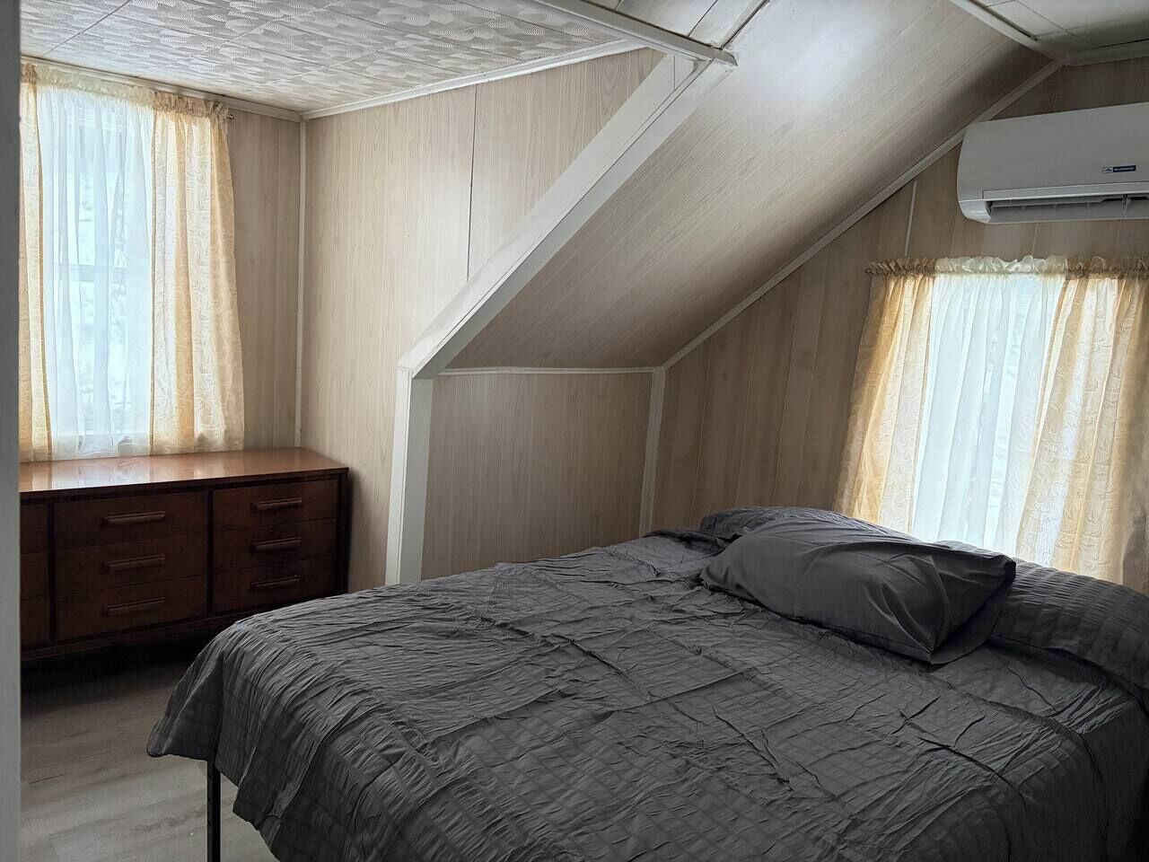 Un dormitorio con techo inclinado, paredes de madera, una cama con ropa de cama gris, un aparador de madera y un equipo de aire acondicionado.
