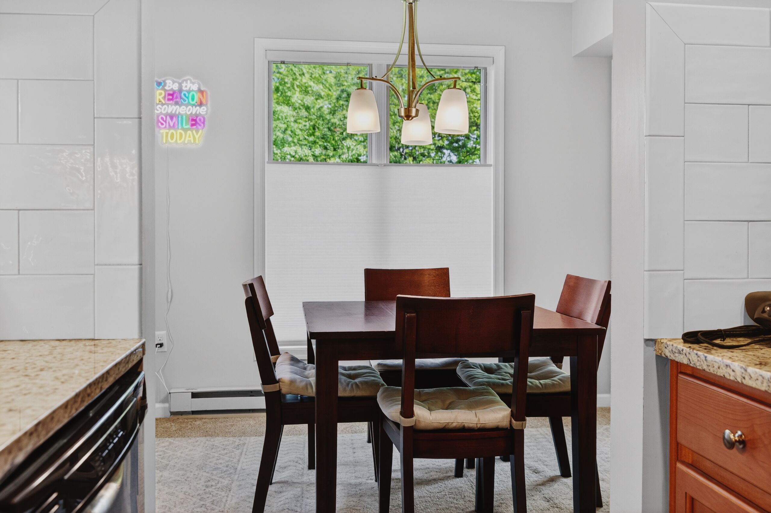 Una cocina con armarios blancos, una mesa de comedor con sillas, una araña y una ventana con follaje en el exterior.