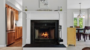 A modern kitchen with a built-in stainless steel refrigerator, wooden cabinets, and a white fireplace with a fire. - Summer Golf - Walk to Ski | Sleeps 12 | Pets OK (Wintergreen Resort)