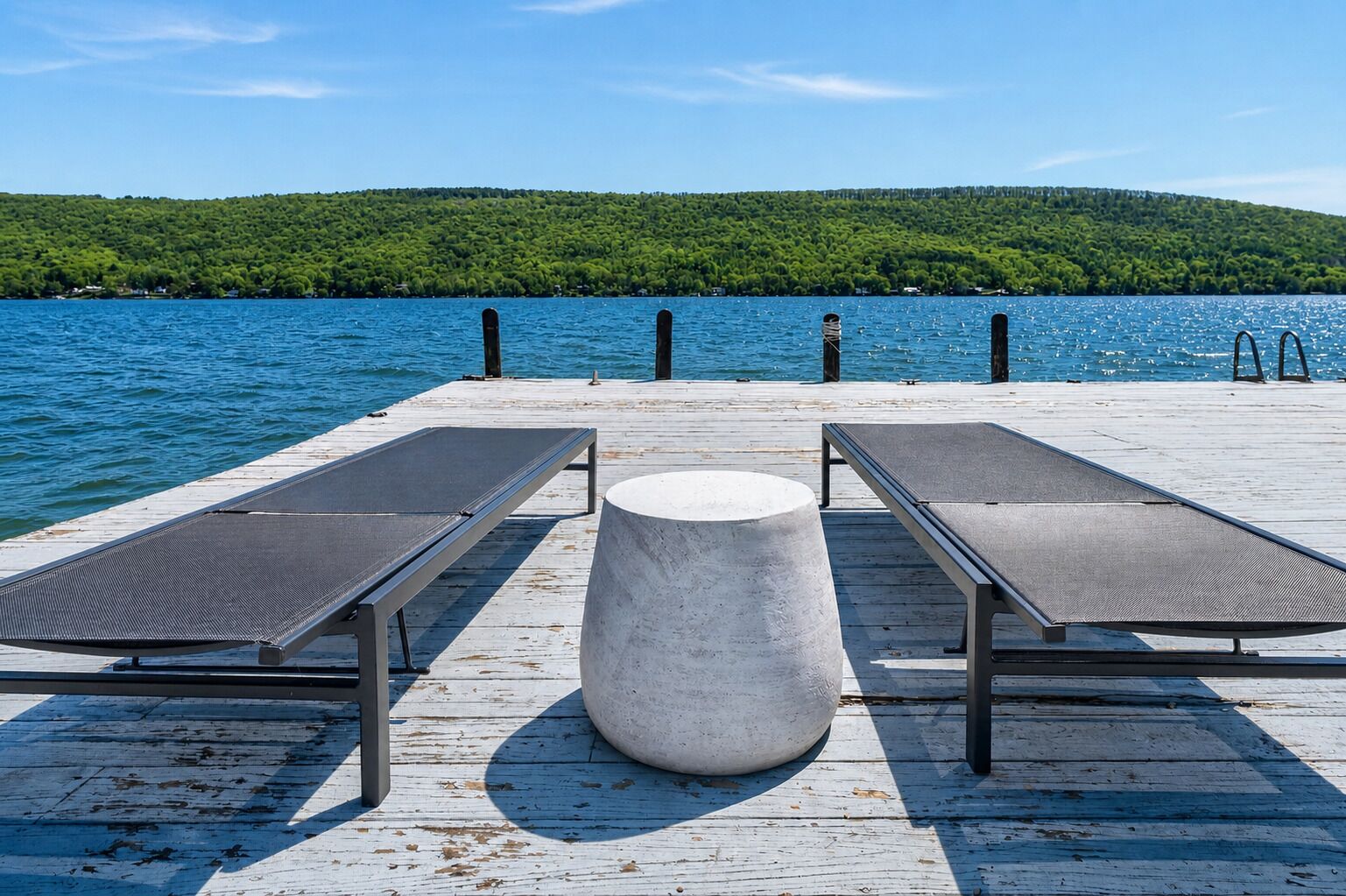 A wooden pier with two sun loungers and a concrete table, overlooking a lake with a forested hill in the background.