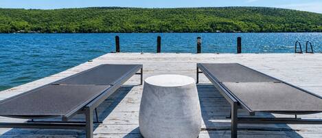A wooden pier with two sun loungers and a concrete table, overlooking a lake with a forested hill in the background.
