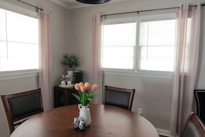 A dining area with a round table, chairs, a vase with flowers, salt and pepper shakers, a small plant, and a window with curtains.