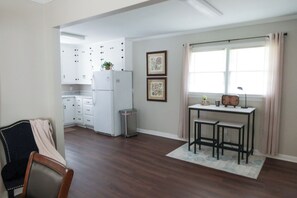 A modern kitchen with white cabinets, a refrigerator, and a trash bin. There is a dining area with a table and chairs, and a window with curtains.