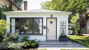 A small, single-story house with a white exterior, a front door, and a large window.