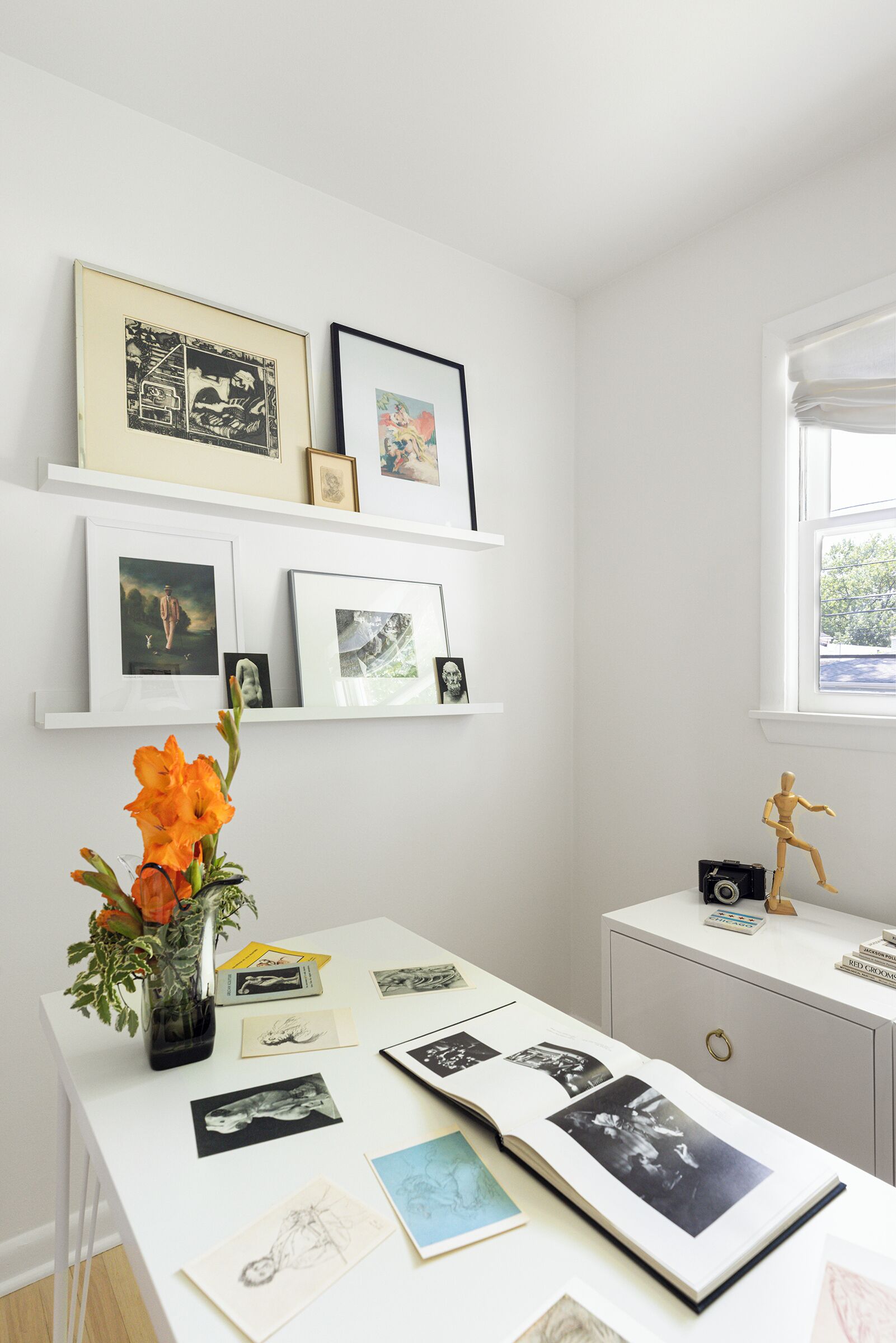 Une pièce avec un bureau blanc, un vase contenant des fleurs oranges, un appareil photo posé sur une petite table et des photos encadrées sur des étagères.