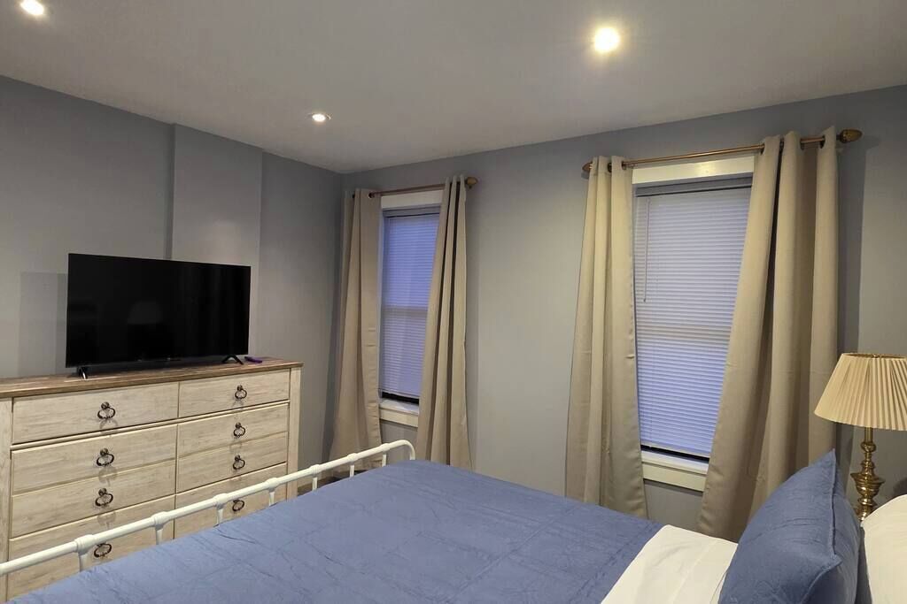 Ein Schlafzimmer mit einem Bett, einem Fernseher auf einer Kommode und zwei mit Vorhängen versehenen Fenstern.