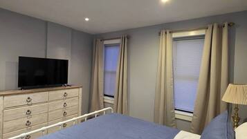 Ein Schlafzimmer mit einem Bett, einem Fernseher auf einer Kommode und zwei mit Vorhängen versehenen Fenstern.