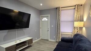A living room with a flat-screen TV, a dark-colored sofa, a white entertainment unit, and a window with patterned curtains. - Work, Crew & Family Stay Near Downtown Reading (Reading)
