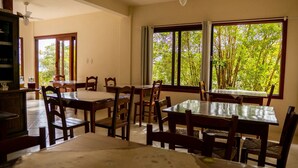 Un espace repas avec des tables et des chaises en bois, de grandes fenêtres donnant sur un paysage verdoyant et un ventilateur de plafond.