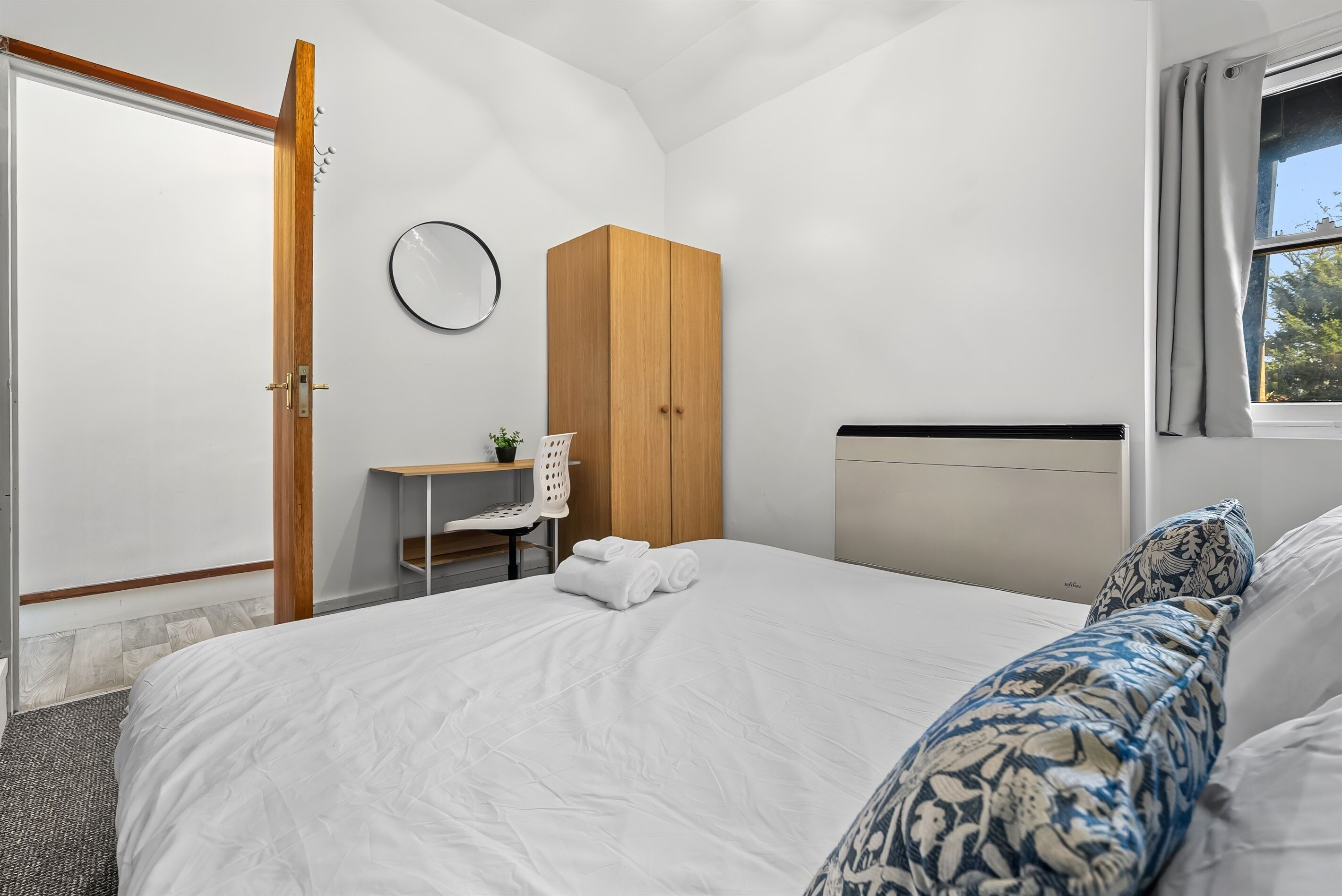 A bedroom with a bed, a wooden wardrobe, a desk with a chair, and a window with curtains.