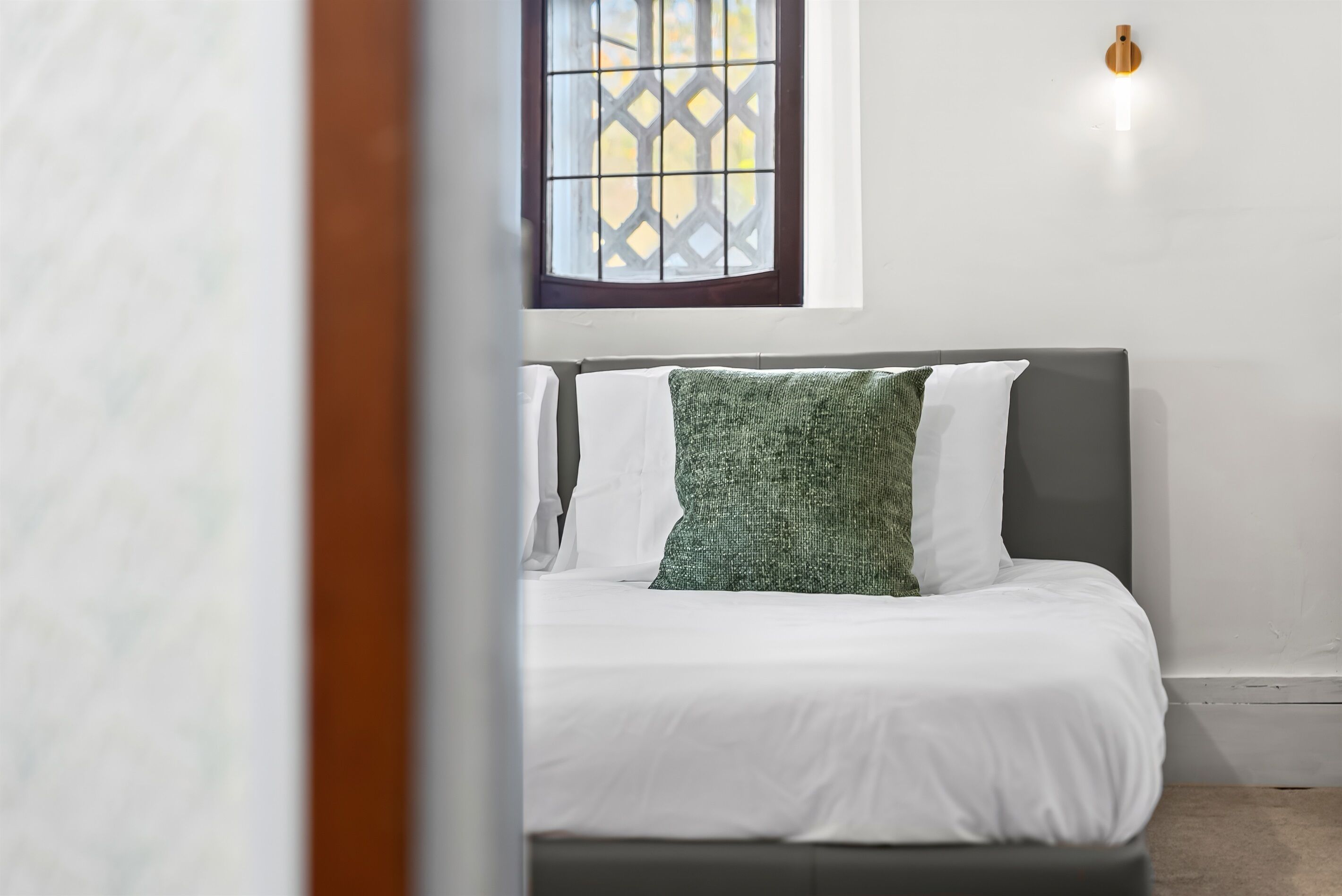 A bedroom with a bed, white bedding, a green pillow, a window with a lattice design, and a wall-mounted light fixture.