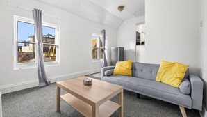 Ein modernes Wohnzimmer mit einem grauen Sofa, gelben Kissen, einem Holztisch und großen Fenstern mit Vorhängen.