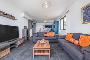 Una sala de estar moderna con un televisor de pantalla plana, una mesa de centro de madera y un sofá gris con cojines naranja.