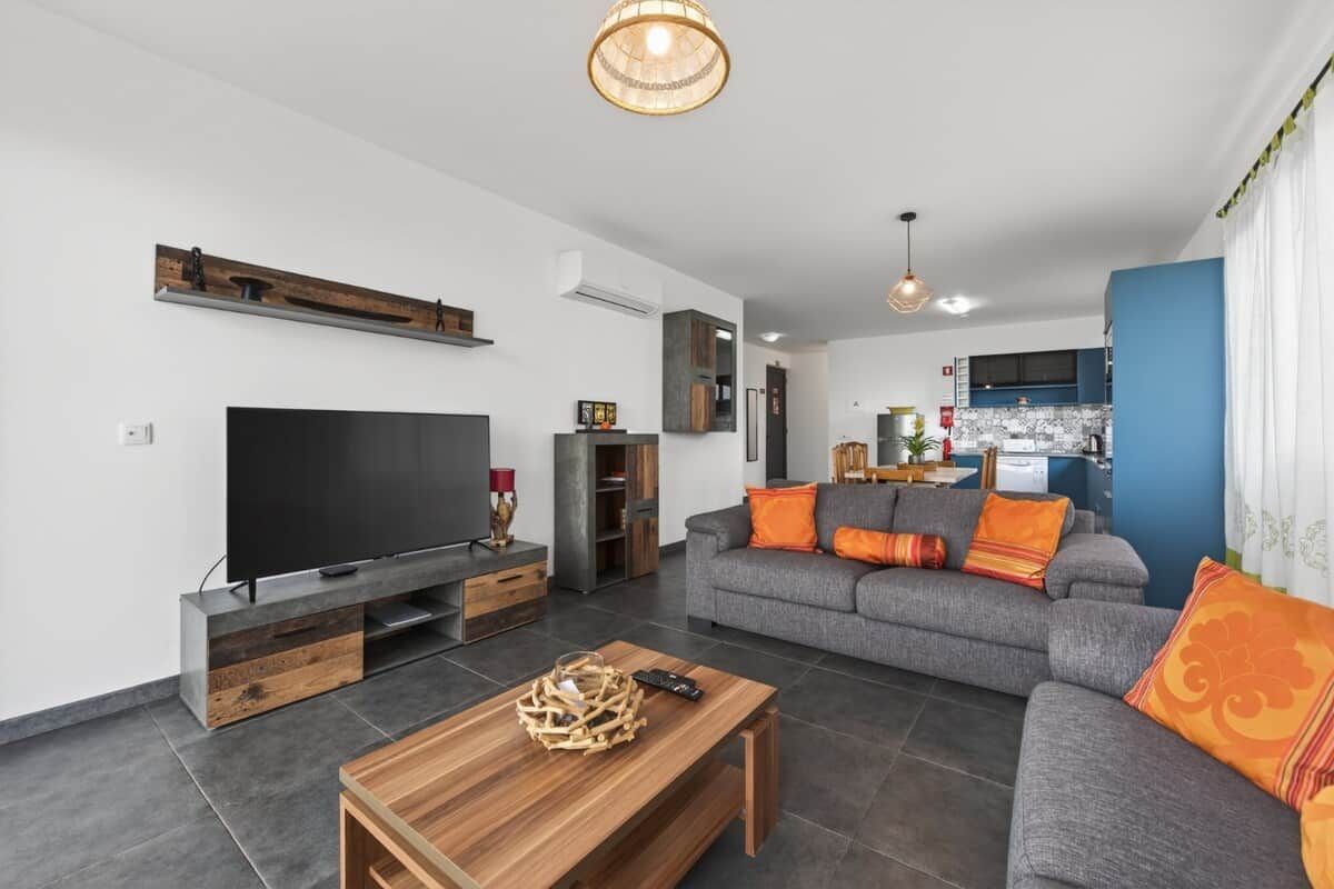 Een moderne woonkamer met een flatscreen televisie, een houten salontafel en een grijze bank met oranje kussens.
