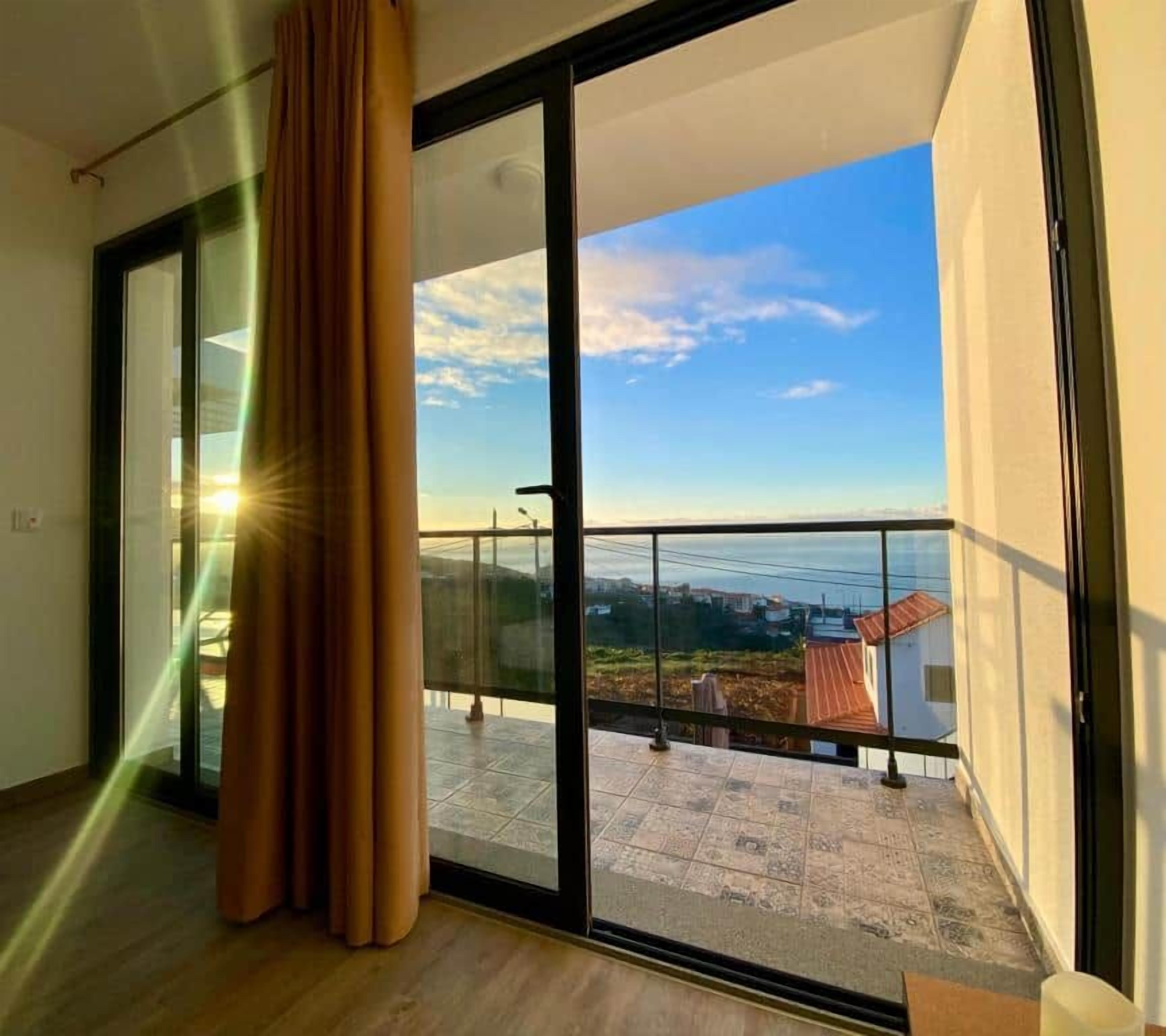 A balcony with a glass door leading to an outdoor terrace, a curtain partially covering the door, and a clear view of the sky and distant buildings.