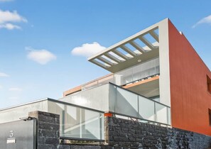 Un edificio moderno con balcón de vidrio, fachada de piedra y cielo azul despejado.