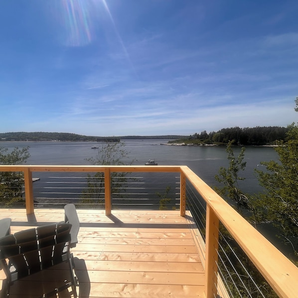A wooden deck with a railing overlooking a calm body of water, boats, and a distant shoreline.