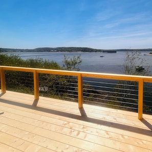 A wooden deck with a railing overlooking a calm body of water, surrounded by greenery and distant hills.