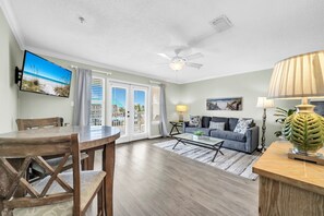 A living room with a television, a ceiling fan, a sofa, a coffee table, and a dining table with chairs. - Cozy Condo – Gulf View & Pool Access • Master bedroom + hallway w/ Bunks (Destin)